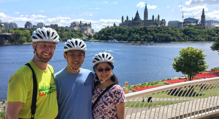 Preferred Partner - Escape Bike Tours & Rentals. Image of a family wearing helmets and standing in front of Ottawa's Parliament buildings. 